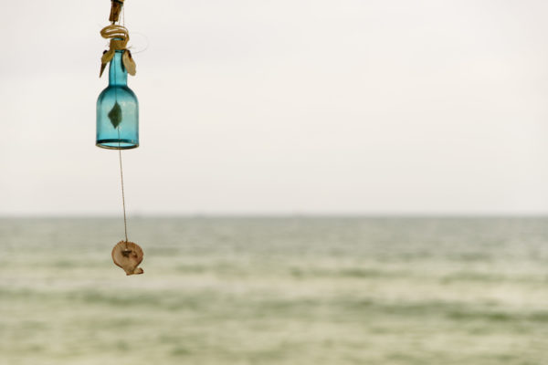 An empty, aqua-blue bottle hanging with a seashell as a summer wind chime decoration and wind chime - shot in an afternoon with sea coast in the background.