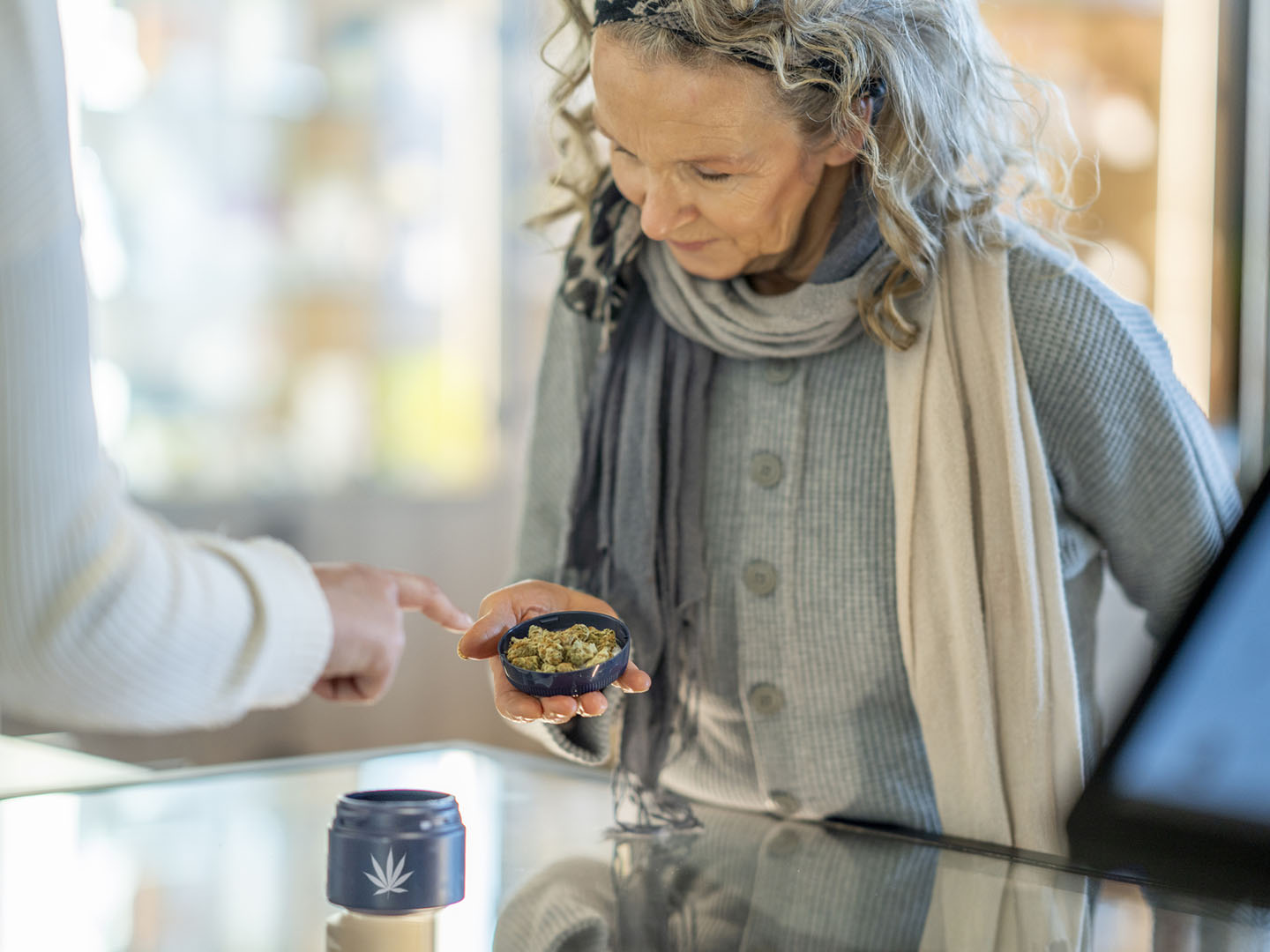 A mature woman stands at the counter of a legal cannabis retailer as she holds out a lid of buds.  She is dressed casually and looking down to inspect the product as the associate tries to sell it to her.